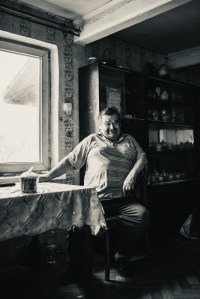 monochrome,image of man in short. sleeved shirt sitting in a kitchen with a drink on the table next to him